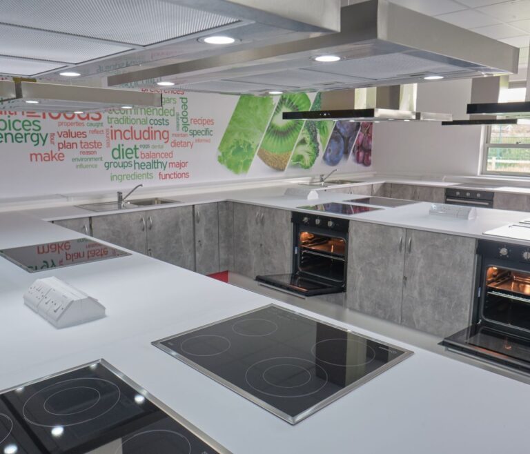 Modern food technology classroom with white worktops, electric hobs, built-in ovens and a colourful food-themed wall display.